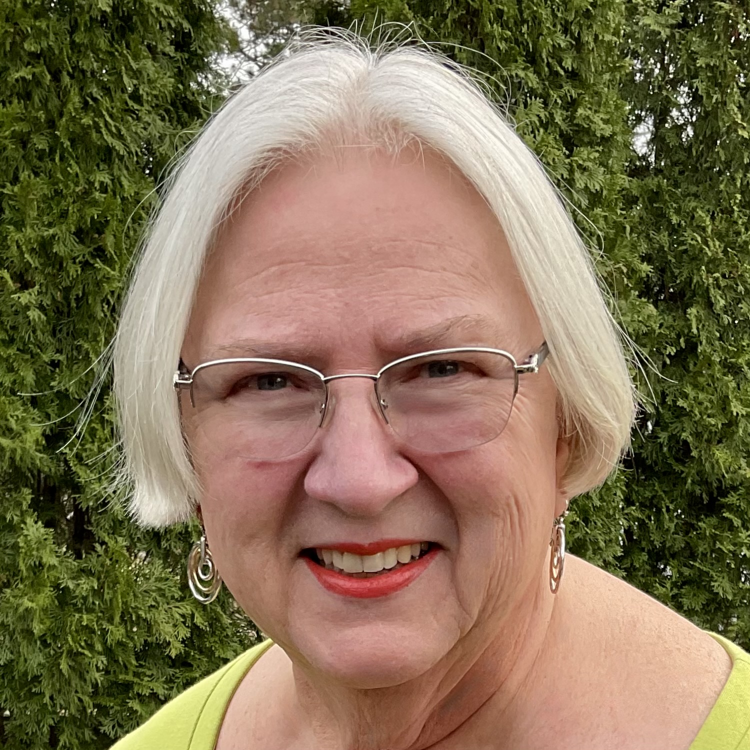 Older woman with short white hair, glasses, and earrings, smiling outdoors in front of green trees.